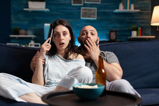 Astonished couple watching movie at night and eating popcorn, drinking beer reacting at film emotion. Focused concentrated confused wife chilling home on confortable couch having facial expression