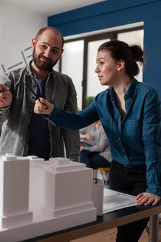 Architect work team talking at professional office standing at desk with maquette building model. Caucasian project partners looking to design blueprint for construction layout