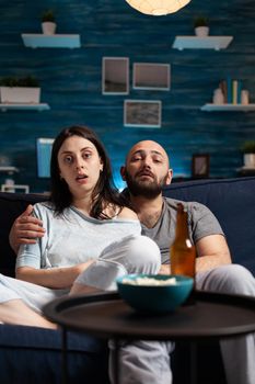 Astonished couple watching movie at night and eating popcorn, drinking beer reacting at film emotion. Focused concentrated confused wife chilling home on confortable couch having facial expression