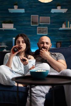 Relaxed excited couple watching TV on couch relaxing late at night laughing having fun drinking beer. People enjoying entertainment show leisure, happiness and married people concept.