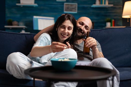 Excited couple in pajamas chilling on couch eating popcorn watching comedy movie, enjoying free time together. Husband and wife relaxing late at night in living room spending time together