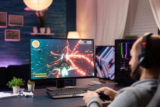 Man with headphones and joystick streaming space shoot videogames in gaming home studio. Professional player man talking with other players online for game competition playing at powerful computer