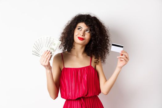 Shopping. Woman thinking and smiling, holding money with plastic credit card, wearing red elegant dress and evening makeup, white background.