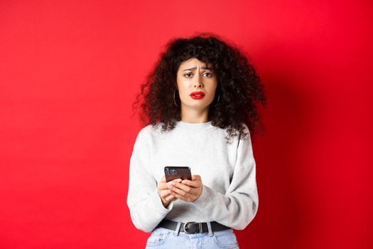 Sad and gloomy woman with curly hair, frowning and feel upset after reading smartphone message, standing disappointed against red background.