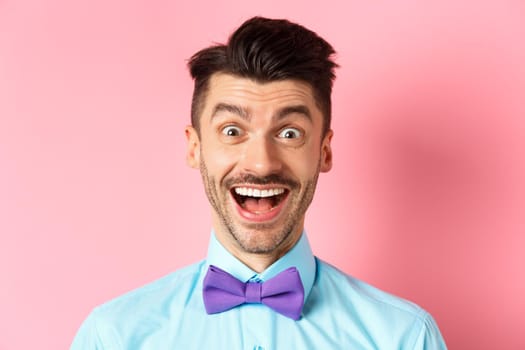 Close-up of surprised and happy young guy, open mouth and smiling amazed, checking out promo offer, standing in bow-tie over pink background.