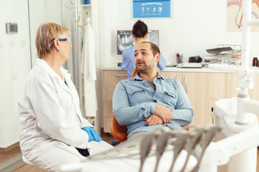 Specialist senior dentist discussing with man patient sitting on dental chair preparing for tooth surgery. In background medical assistant looking into monitor examining stomatological radiography