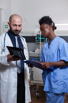 Physician doctor with afro american assistant monitoring recovery treatment using tablet computer working in hospital ward. Patient woman lying in clinical bed discussing with specialist surgeon