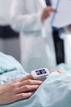 Close up of hospitalized woman with finger heart rate monitor showing pulse while sitting in bed during cardiology examination in hospital ward. Patient with oximeter waiting for illness treatment