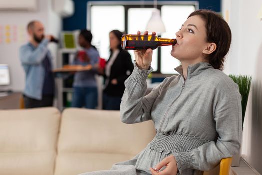 Cheerful woman drinking from bottle of beer after work at office with colleagues. Coworkers meeting at party with food and drinks to celebrate break together. Fun activities enjoyment