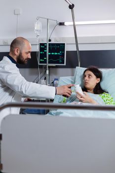 Man doctor monitoring sick patient during medical appointment explaining pill treatment against pain in hospital ward. Patient resting in bed with nasal oxygen tube discussing pharmacy medication