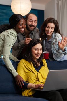 Multi ethnic friends waving at laptop webcam during video call sitting on couch late at night. Group of multiracial people spending time together sitting on couch late at night in living room.