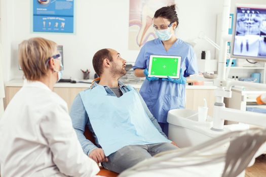 Nurse pointing at green screen display while patient sitting on dental chair. Senior doctor explaining using mock up green screen chroma key tablet with isolated display working in hospital office