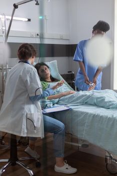 Medical team checking sick woman analyzing disease symptom during recovery appointment in hospital ward. Patient resting in bed discussing illness treatment while doctor writing expertise on clipboard