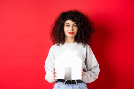 Real estate. Young caucasian woman in casual clothes showing paper house cutout, buying property or renting apartment, standing on red background.