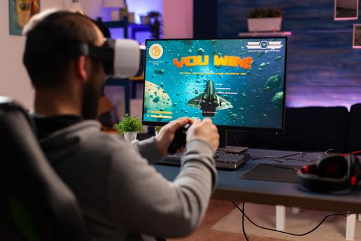 Gamer man winning space shooter game use virtual reality goggles. Competitive player using joystick for online championship sitting on gaming chair late at night playing on professional computer