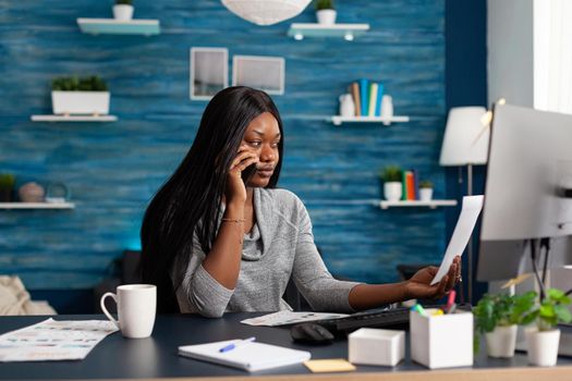 Black sad student interacting with collegue explaining communication course ideas using phone sitting at desk in living room. African woman working at academic homework using elearning platform