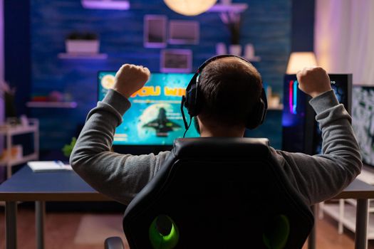 Back view of happy gamer playing and winning space shooter game in gaming home room. Cyber performing on powerful computer streaming videogames using professional headphones for online championship