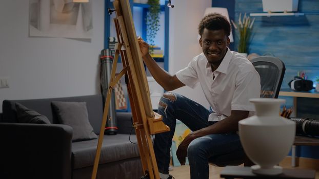 Black young adult sitting in artwork studio drawing vase with pencil and white canvas on easel. African american man with creative occupation making innovative masterpiece and fine art