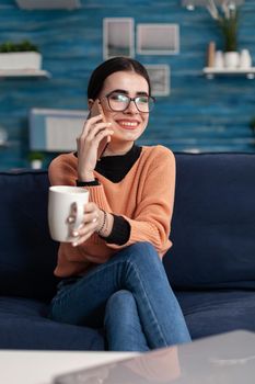 Portrait of teenager talking about lifestyle on modern smartphone while laughing with her friend sitting on couch in living room. Young woman having fun during funny lifestyle conversation call