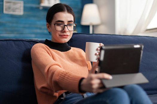 Student reading online book on digital application using tablet computer, sitting on sofa in living room. Teenager studying social communication using university mobile app drinking cup of coffee