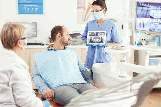 Medical nurse showing on tablet teeth x-ray showing it with patient. Dentist senior doctor and nurse working together in modern stomatological clinic, explaining treatment to man patient