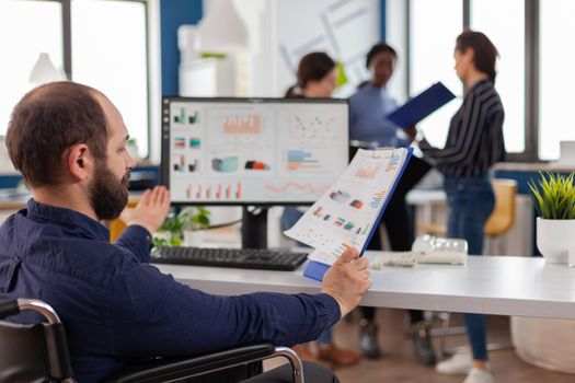 Paralized, immobilized, handicapped marketing manager project sitting in wheelchair in start up business office reading checking reports analysing datas. Diverse team discussing financial project.