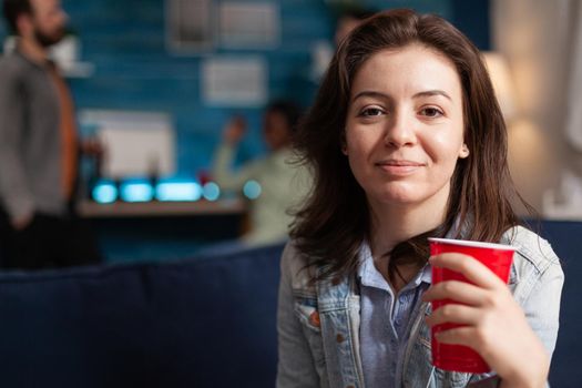 Portrait of beautiful woman holding beer glass looking into camera during night party. Multi-ethnic diverse group of friends gathered together to celebrate birthday late at night cheerful young people