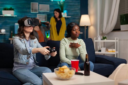 Mixed race friends having online virtual games competition wearing vr headset, using wireless controller, hanging out late in the evening sitting on sofa drinking beer and enjoying snacks.