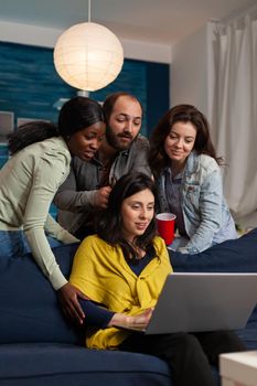 Multi ethnic friends hanging out talking on webcam laptop during video call. Group of multiracial people spending time together sitting on couch late at night in living room.