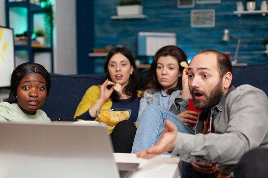 Multiracial friends relaxing on couch while watching comedy looking shocked during movie night. Group of multi-ethnic people spending time together drinking beer, eating snacks late at night.