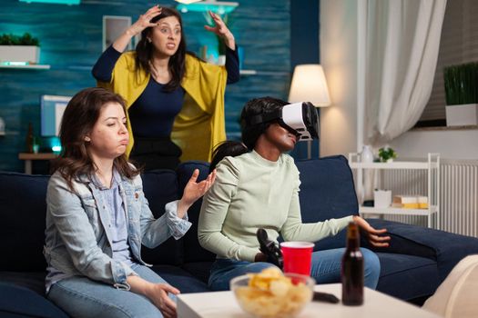 Upset multi ethnic women after losing while playing video games wearing virtual reality goggles. Mixed race group of people hanging out together having fun late at night in living room.