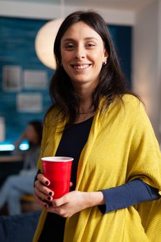 Portrait of cheerful woman looking into camera while in background her friends having fun celebrating birthday party late at night in living room. Group of multiethnic people drinking beer and talking.