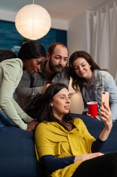 Multi ethnic friends bonding and talking on smartphone during video call in home living room. Group of multiracial people spending time together sitting on couch late at night in living room.