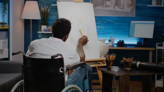 Disabled african american artist working on drawing with pencil on white canvas in workshop space. Invalid black young person sitting in wheelchair creating authentic fine art of vase