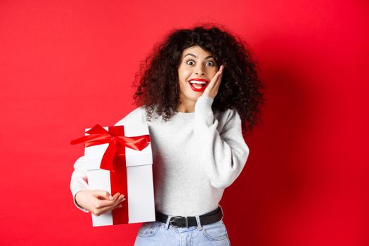 Surprised pretty girl receive Valentines day romantic gift, holding present box and looking with disbelief and happiness at camera, standing on red background.