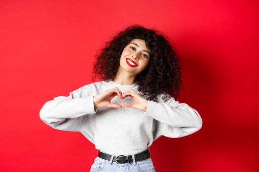 Beautiful young woman with curly hair showing heart gesture, say I love you and smile romantic at camera, standing on red background.