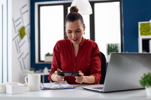 Businesswoman sitting at desk in corporate office playing video games, using smartphone. Entrepeneur taking a break from wwork having fun with games holding phone. Employer having fun at wwork.