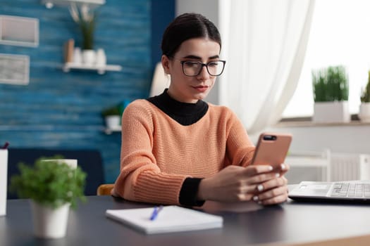 Student looking at smartphone screen while texting message sitting at desk table in living room. Student browsing lifestyle information on internet during coronavirus quarantine