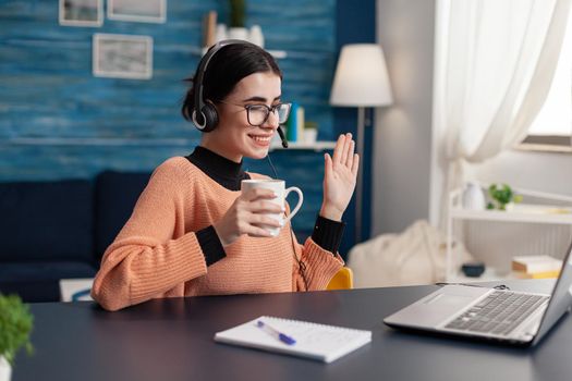 Student wearing headphone while waving her college treacher during online videocall meeting. Woman holding cup of coffee looking at laptop computer sitting at desk in living room