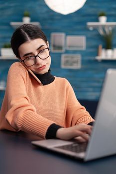 Student browsing information on laptop computer for digital seminar. Teenager woman speaking at phone whit collegue about online university meeting while sitting at desk in living room