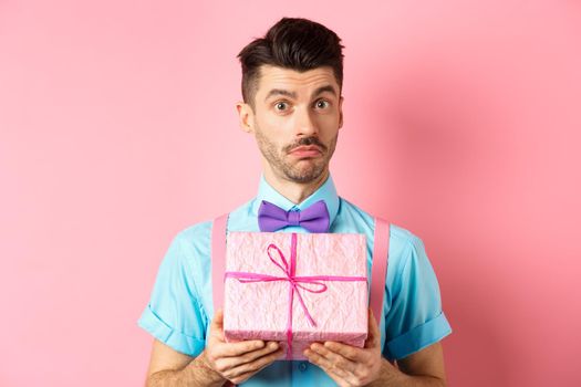 Holidays and celebration concept. Attractive young man with moustache, wearing festive outfit with bow-tie, showing cute gift box and looking at camera, standing over pink background.