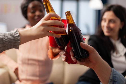 Close up of bottles and cups of beer from cheerful friends after work at office party. Multi ethnic happy group cheers for celebrating entertainment indoors with snacks drinks food