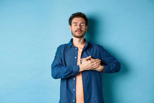 Romantic and nostalgic guy holding hands on heart with eyes closed, keep warm memories, standing on blue background.