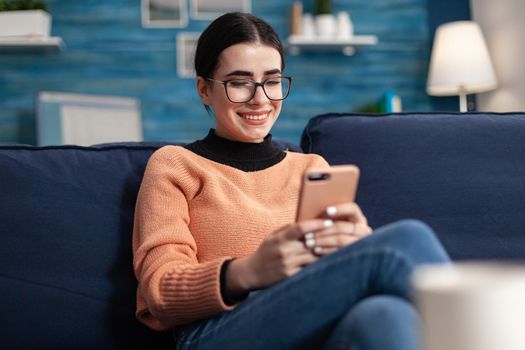 Relaxed student sitting on couch in living room while writing message holding modern smarphone in hands. Teenager woman chatting on social network looking at phone screen