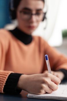 Selective focus on student hand taking notes on notebook for university exam before online meeting on zoom. Young woman with headphone doing homework while sitting at desk table in living room