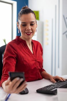 Business woman sitting at desk working on financial charts, checking and looking at phone. Executive reading text from smartphone about project details. Mobile device used by entrepreneur.