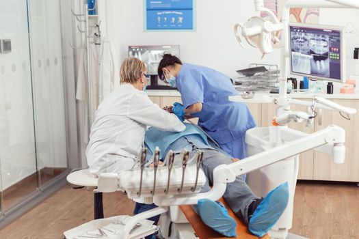 Senior dentist doctor and nurse preparing patient for removing crowns. Medic orthodontist wearing protection mask treating teeth of sick man patient in stomatological office sitting on chair