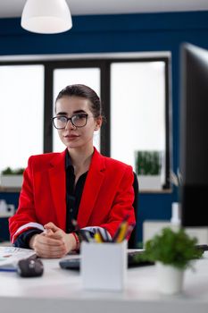 Portraif of confident businesswoman sitting at desk in corporate office workplace building, with finger crossed wearing glasses looking at camera. Financial charts and graphs.