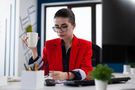 Entrepreneur holding cup of coffee in business workplace office, cheking notes about financial expertise on clipboard. Manager sitting at desk table reviewing strategy. Caffeine addicted.