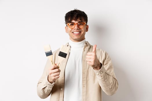 Handsome natural guy in glasses recommending store, showing painting brushes for renovation and decor, make thumbs up in approval, standing on white background.